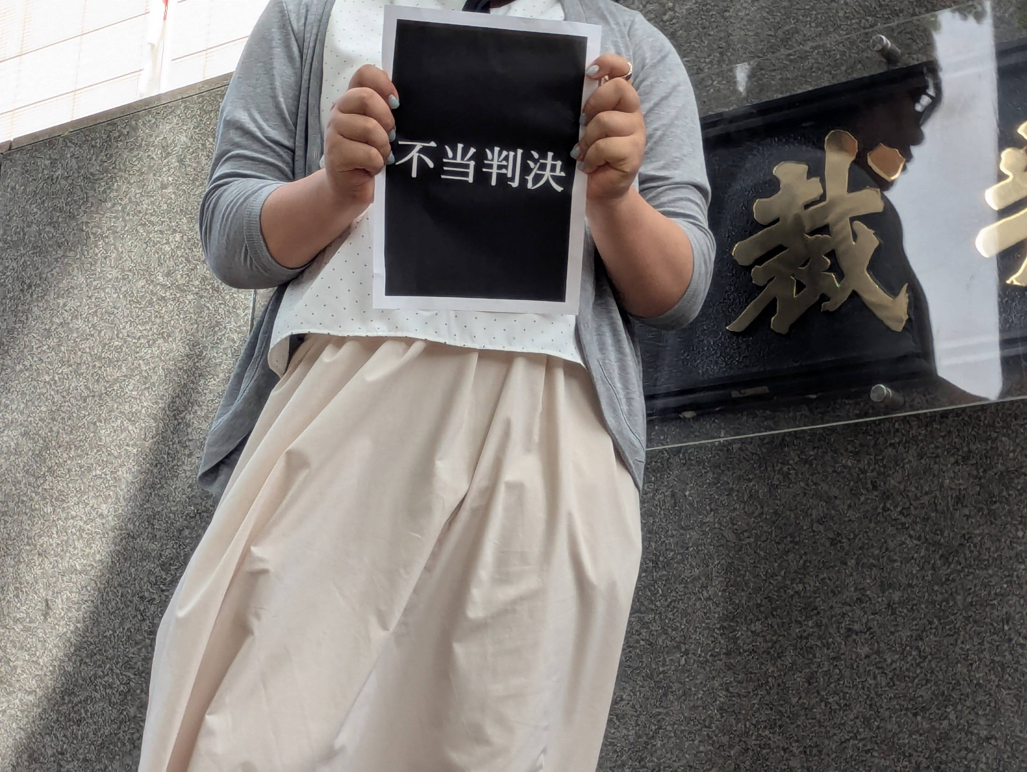 原告人が、黒い紙に白い文字で「不当判決」と書かれた看板を持っている。背景には灰色の壁と「裁判所」と刻まれた金のプレートがある