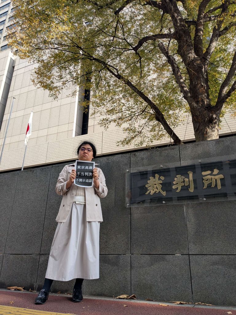 控訴人が、黒い紙に白い文字で「東京高裁不当判決。不開示追認」と書かれた看板を持っている。背景には灰色の壁と「裁判所」と刻まれた金のプレートがあり、壁の奥に日本国旗と裁判所建物がある