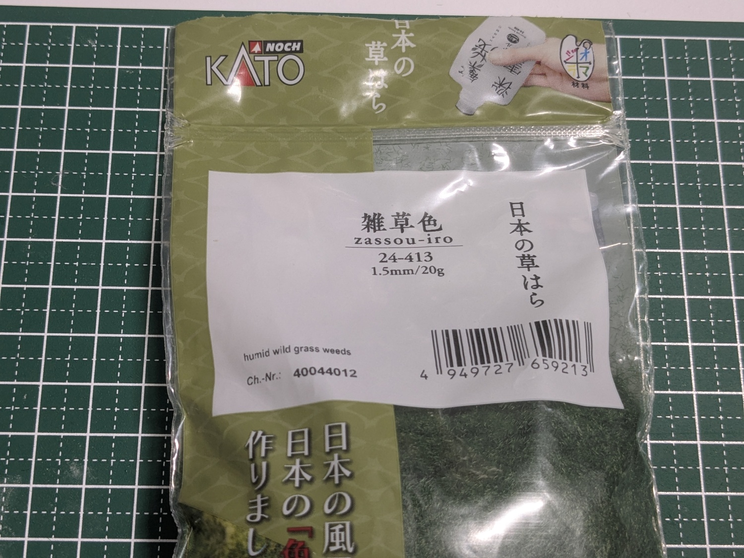 KATOの情景素材、日本の草はらのパッケージ外観写真。雑草色、型番24-413が印字されている。包装は右半分が透明で内容物の様子が見える。濃い緑色の細い繊維が大量に入っている